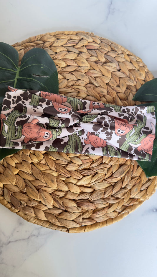 A twist headband on a wicker tray with monstera leaves as photo props. This headband has a western vibe with its fluffy highland cows, green cactuses, and cow print in the background.