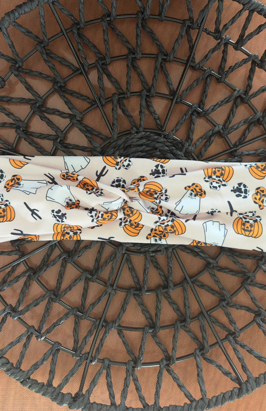 A twist headband on a black wicker tray on top of orange tulle fabric. The headband features adorable ghosts and pumpkins with cowprint cowboy hats on. There are black cactus's too