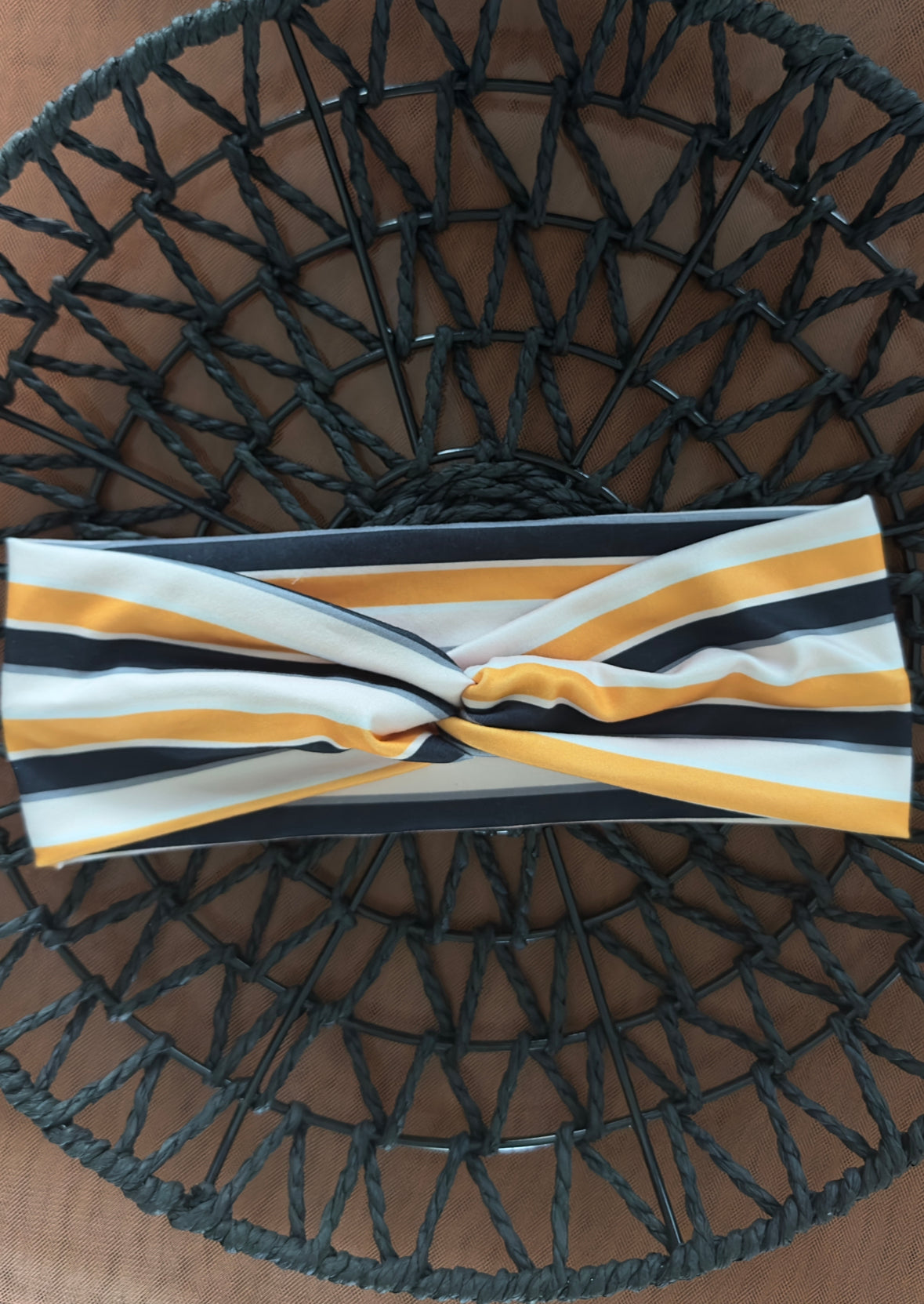 A twist headband on a black wicker tray on top of orange tulle. The headband is striped in the colors White, orange, black. The stripes are hortizontal.