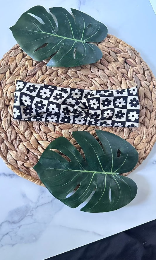 Black and white checkered floral twist headband on a whicker placemat with monstera leafs on a marble background.
